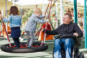 man in a wheelchair smiling and helping two toddlers playing on a playscape